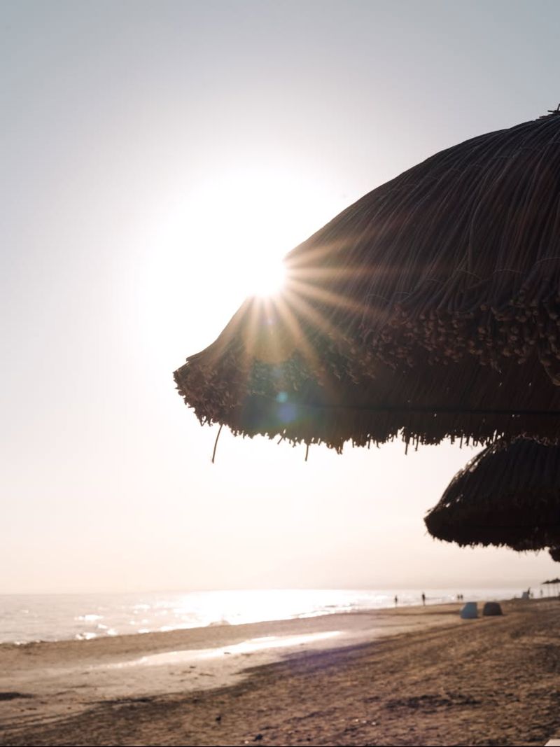 The sun low behind a thatched parasol on the shore