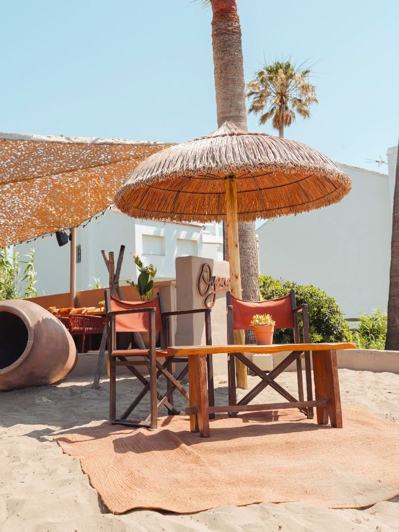 Director’s chairs and a wooden table on a rug in the sand beneath a parasol