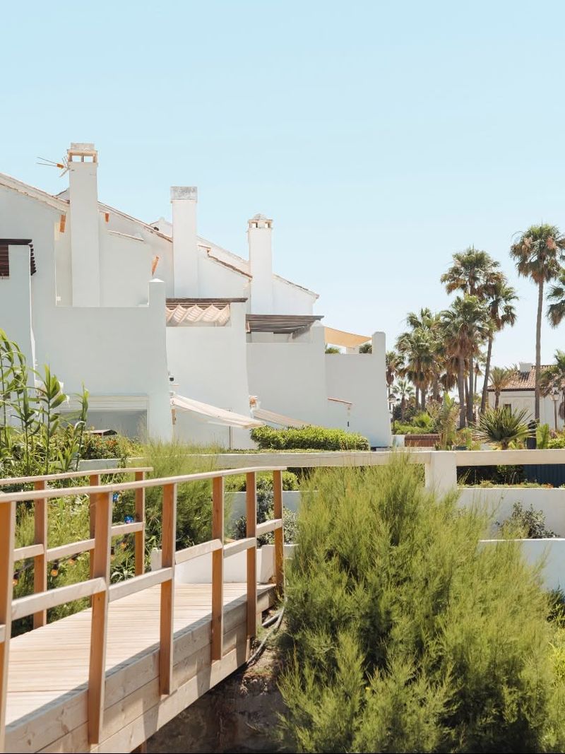 A wooden boardwalk leading past whitewashed houses and palms towards the beach