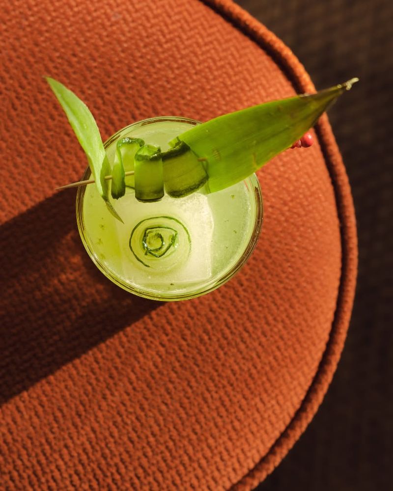 A pale green cocktail with a leaf garnish, seen from above on a terracotta cushion