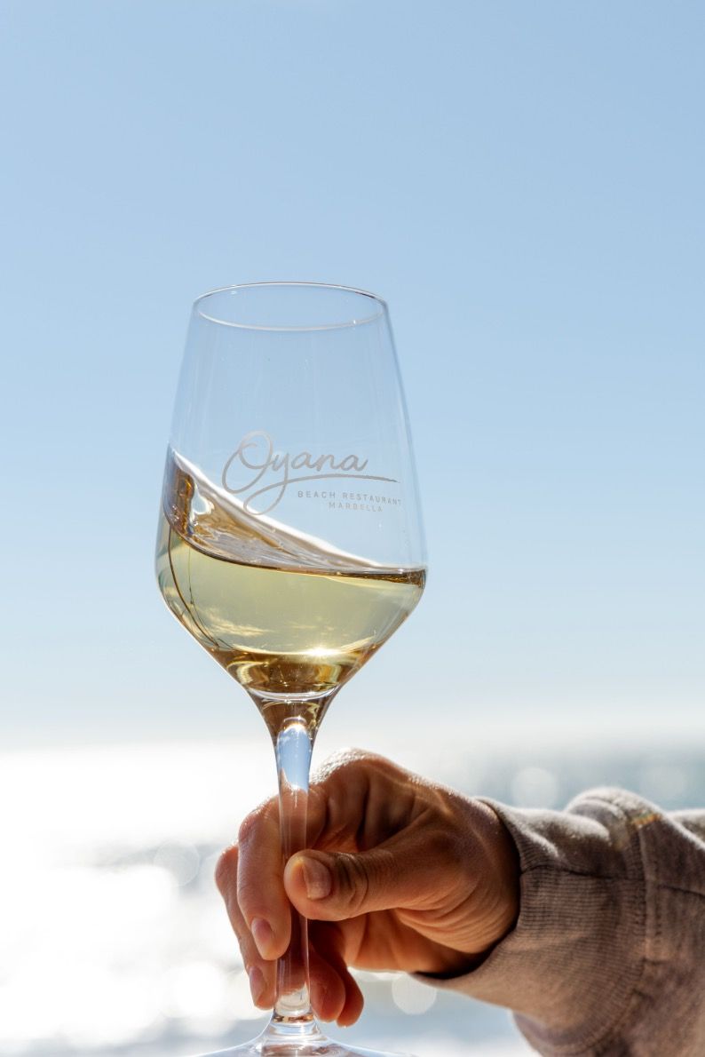 A hand raising a glass of white wine etched with the Oyana logo against the sea
