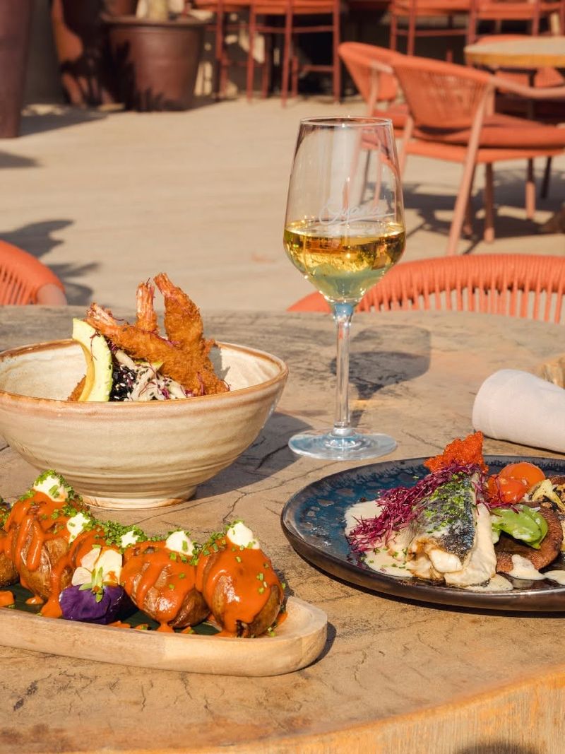Sharing plates and a glass of white wine on a terrace table, with coral chairs behind