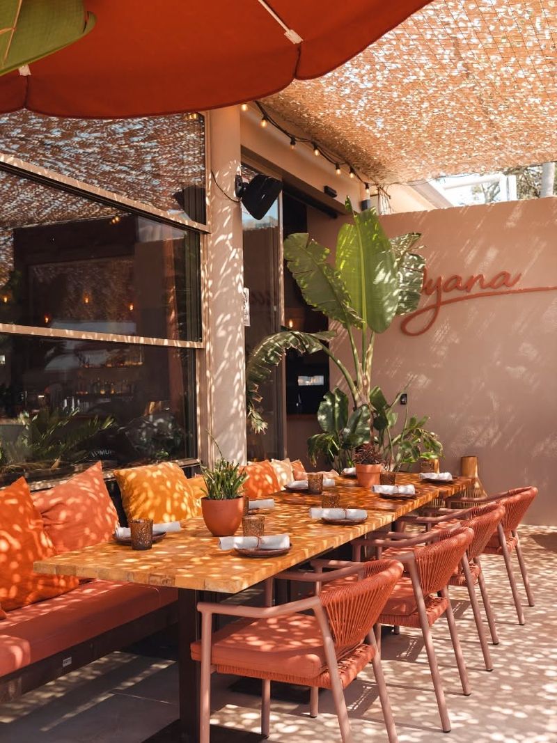 Long terrace table with coral cushions under a shaded canopy and the Oyana sign on the wall
